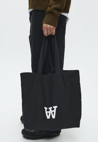 Schwarze Tote-Bag aus Canvas mit kontrastierendem weißen Logo, ausgestattet mit robusten Griffen und einem minimalistischen Design, das von einer Person gehalten wird.