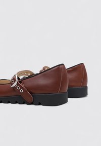 Zapatos de cuero marrón con una suela negra plana y texturizada, que cuentan con una correa con un hebilla de plata y perforaciones a lo largo de los lados.