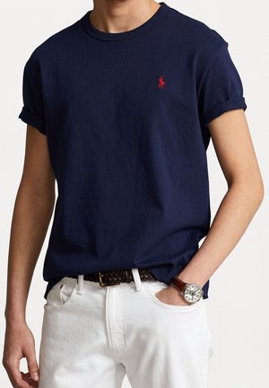 Navyblauwe katoenen t-shirt met opgerolde mouwen, voorzien van een klein rood logo op de borst. Draag het met witte broek en een bruine gevlochten riem.