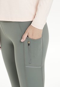 Vaaleanpunainen pitkähihainen paita, yhdistettynä vihreisiin, muotoilultaan ihonmyötäisiin legginsseihin, joissa on vetoketjullinen sivutasku ja kontrastiväriset ompeleet.