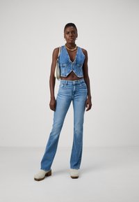 Denim cropped vest met een diepe V-hals, gecombineerd met lichtblauwe uitlopende jeans. Witte platformklompen geven extra hoogte, aangevuld met een klein tasje.