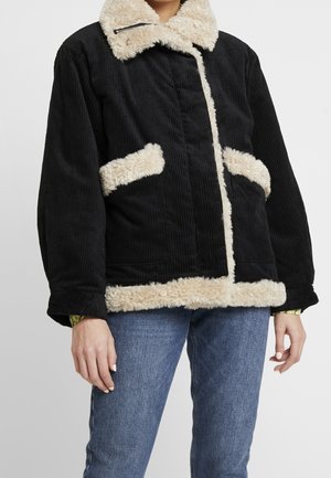 Giacca in velluto a coste nera con fodera in shearling color crema su colletto, tasche e orlo, indossata con jeans blu.