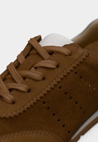 Gros plan d'une basket en suède marron avec des lacets assortis, une couture en zigzag, un côté perforé et un détail blanc sur la languette.