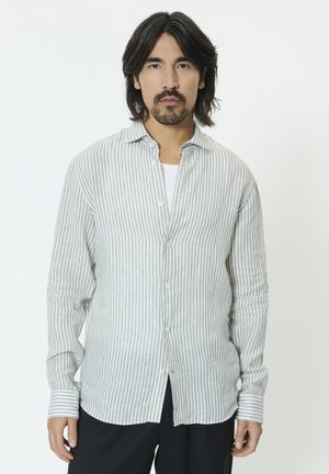 Man met schouderlang haar en baard, gekleed in een lichtgrijs-witte verticale gestreepte overhemd over een wit ondershirt, staand tegen een witte achtergrond.