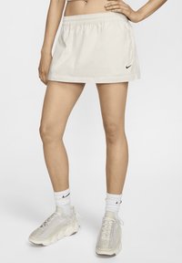 Világos bézs Nike szoknya, rugalmas derékkal, minimalista dizájnnal és egy kicsi logóval az oldalon. Fehér zoknikkal és sportcipőkkel viselve.