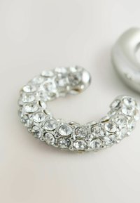 Sølvfarvet smykke formet som en halvmåne, indlagt med mange klare funkende ædelsten, med en glat metallisk finish.