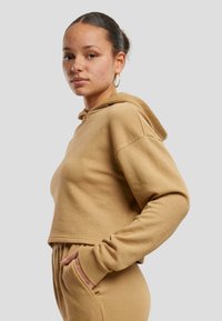 Sudadera con capucha recortada en tela de algodón color beige, con un bolsillo delantero, puños acanalados y texturizada suave. Combinada con pantalones a juego.