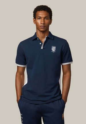 Homme debout les mains dans les poches, portant un polo bleu marine et un pantalon assorti avec les logos du Hackett London Rowing Club.