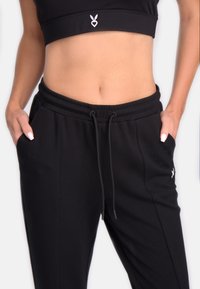 Pantaloni de sport negri cu talie elastică, buzunare laterale și șnur. Prezintă un mic logo alb pe coapsa stângă.