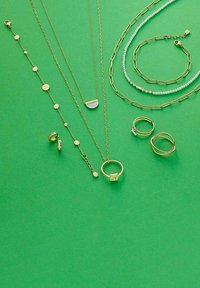 Gouden sieraden tentoongesteld op een groene achtergrond, inclusief gelaagde kettingen, ringen en armbanden met verschillende texturen en vormen.