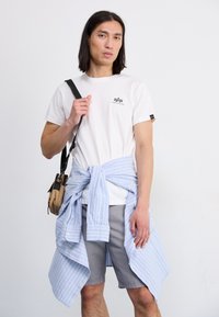Weißes T-Shirt mit einem Logo, graue Shorts und ein blau gestreiftes Hemd, das um die Taille gebunden ist. Braune Umhängetasche mit schwarzen Riemen.