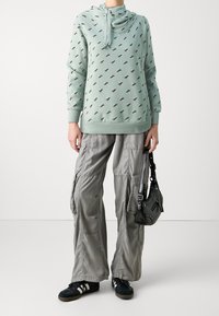 Sudadera con capucha verde menta con un patrón geométrico, combinada con pantalones cargo gris claro y zapatillas negras. Bolsa negra con detalles metálicos.