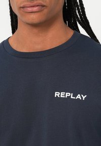 Gros plan d’une personne portant un T-shirt bleu marine avec le mot "REPLAY" imprimé en blanc sur la poitrine.