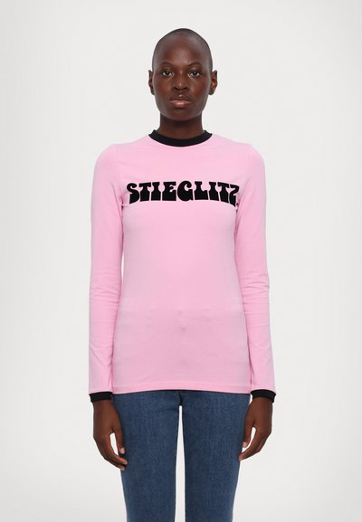 Lichtroze shirt met lange mouwen en zwarte halslijn en manchet; heeft opvallende zwarte tekst "STIEGLITZ" over de voorkant. Glad katoenmateriaal.