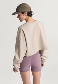 Μπεζ cropped φούτερ με φαρδιές ραχιαίες ραφές, συνδυασμένο με ανοιχτό μοβ σορτς με λείο ύφασμα και ελάχιστες ραφές. Καπέλο σε ελιά χρώμα.