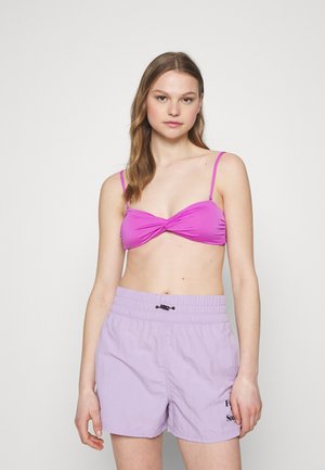 Rožinė bikini viršutinė dalis su sukimo detale ir plonomis petnešėlėmis, derinama su laisvomis levandų maudymosi šortais, kurie turi elastingą juosmenį ir raištelį.