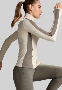 Langärmliges Sportoberteil in hellem Creme und Taupe mit hohem Kragen und seitlichen Einsätzen; kombiniert mit enganliegenden Leggings in Olivgrün.
