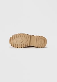 Suela de caucho beige de una bota con surcos profundos, patrones de tracción en forma de diamante y un logo circular cerca del talón sobre un fondo blanco.