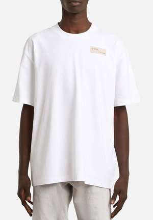 Personne à la peau foncée portant un T-shirt blanc surdimensionné avec une petite étiquette rectangulaire beige sur la partie supérieure gauche de la poitrine et un pantalon gris clair.