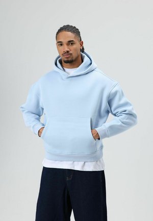 Lichtblauwe hoodie gemaakt van een zachte katoenmix. Voorzien van een voorzak, geribbelde manchetten en een capuchon met trekkoord. Draagt een witte shirt eronder.
