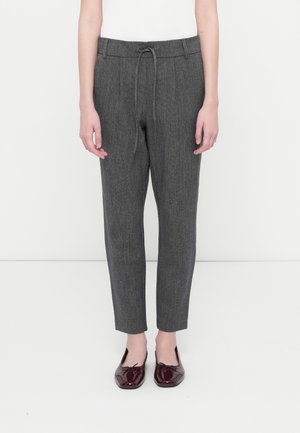 Slim-fit grijze broek met aantrekkoord in de taille, gedragen met bordeauxrood lakleren ballerina's door een persoon die tegen een witte achtergrond staat.