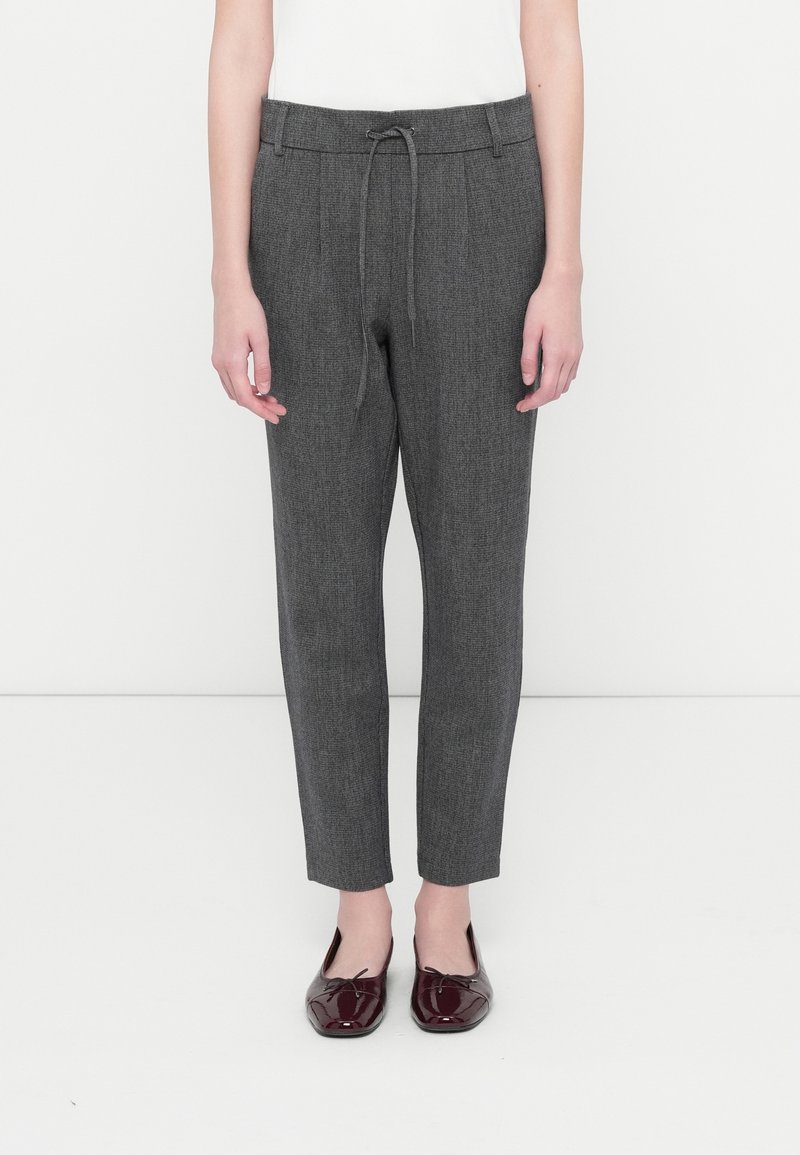 Pantalon gris ajusté avec taille à cordon, porté avec des ballerines en cuir verni bordeaux sur une personne debout devant un fond blanc.