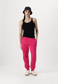 Črn prilegajoč tank top, svetlo roza trenirke z bočnimi žepi in bele športne copate z bež podplatom. Model nosi mornarsko modro klobuk.