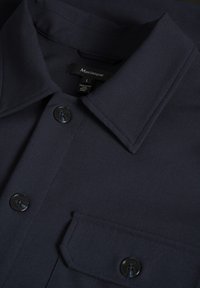 Gros plan d’un col de chemise bleu marine foncé avec des boutons noirs et une poche poitrine, montrant l’étiquette de la marque Matinique à l’intérieur du col.
