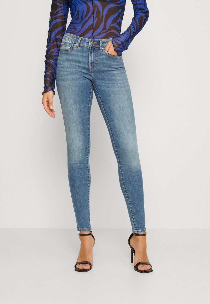 Skinny blue jeans with a faded finish, featuring five pockets, belt loops, and button closure, paired with a patterned long-sleeve top.