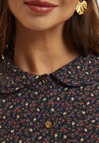 Bluse mit Blumenmuster auf dunkelblauem Hintergrund, Rüschensaum am Kragen und goldfarbener Knopfdetaillierung. Verziert mit kleinen Blumen in Rot, Blau und Creme.