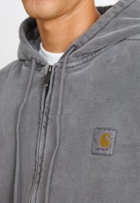 Grauer Reißverschluss-Hoodie aus weichem Stoff, mit Kängurutasche, Kapuze mit Kordelzug und kleinem gesticktem Logo auf der linken Brust.