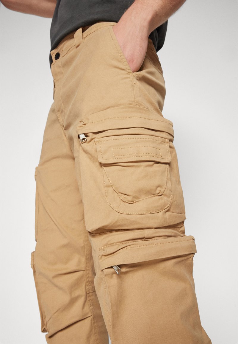 Beige Cargo-Hosen mit mehreren Taschen, Reißverschluss-Details und einer lässigen Passform. Hergestellt aus strapazierfähigem Baumwollmaterial mit einer glatten Textur.
