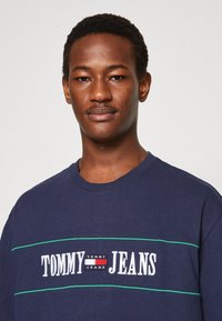 T-shirt in cotone blu navy con collo rotondo, caratterizzato da testo ricamato bianco "TOMMY JEANS" e strisce verdi orizzontali sul petto.