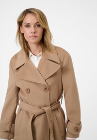Beige double-breasted coat with large lapels, button accents, and a tied waist. Made from textured fabric, featuring cuff details.