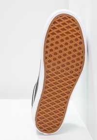 Semelle de chaussure avec motif gaufré marron en caoutchouc et semelle intermédiaire blanche, positionnée verticalement contre un fond blanc d'angle.