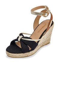 Black fabric wedge sandal with gold ankle strap and knot detail. Espadrille-style sole with woven texture, featuring a tan interior.