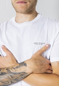 Tricou alb din bumbac care prezintă un mic logo negru cu textul "ARMANI EXCHANGE MILANO/NEW YORK" în zona pieptului stâng.