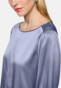 Silky, light blue blouse with a round neckline, smooth texture, and contrast dark shoulder accents, featuring long, loose-fitting sleeves.