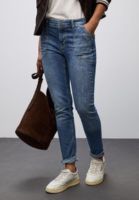 Pantalones vaqueros azules de corte Slim con puños remangados, que cuentan con bolsillos frontales y un ligero desgaste. Combinados con zapatillas blancas y un bolso de ante marrón.