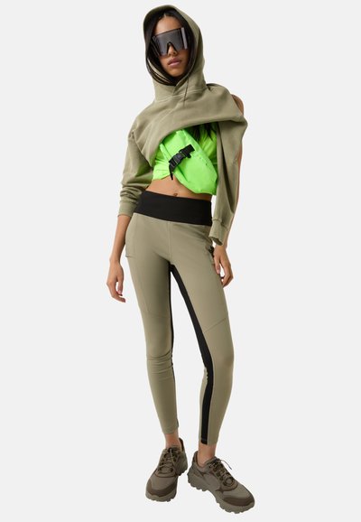 Olive green hooded sweatshirt, bright green cropped top with buckle, beige leggings with black side panel, and chunky brown sneakers.