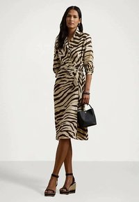 Vrouw in een kokerjurk met zebra-print tot boven de knie, zwarte wedge-sandalen, met een kleine zwarte handtas tegen een effen achtergrond.