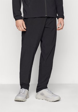 Mand iført sort jakke med lynlås, sorte bukser og lysegrå sportssko, stående på en hvid gulv mod en hvid baggrund.