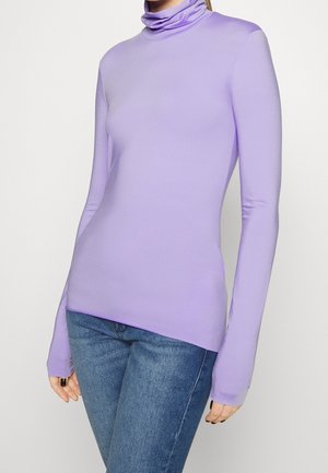 Femme portant un haut moulant à col roulé lavande à manches longues associé à un jean bleu sur un fond uni.
