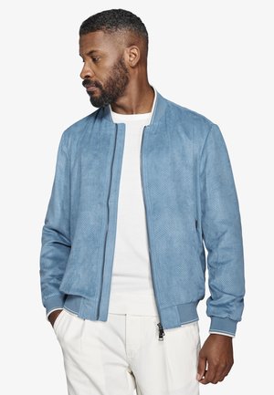 Mężczyzna z krótką brodą, ubrany w jasnoniebieską, perforowaną kurtkę bomberkę na białym swetrze i białych spodniach, patrzący w lewo.