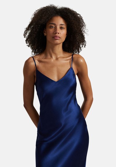 Vrouw met krullend haar, gekleed in een mouwloze, marineblauwe satijnen jurk met dunne bandjes, staand tegen een effen witte achtergrond.