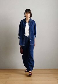 Τζιν πουκάμισο με γυρισμένα μανίκια και κουμπωμένο μπροστά; συνδυασμένο με αντίστοιχα τζιν παντελόνια. Λευκό πουκάμισο από μέσα, κόκκινη τσάντα και κόκκινα παπούτσια.