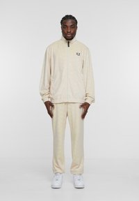 Beige fleece trackjacket with zip, side pockets, and logo, combined with matching pants. Textured fabric, relaxed fit, complemented by white sneakers.