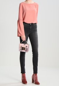 Blouse rose clair à manches évasées, jean skinny noir et bottines bordeaux à talons. Porte un sac à main rose avec des détails floraux.