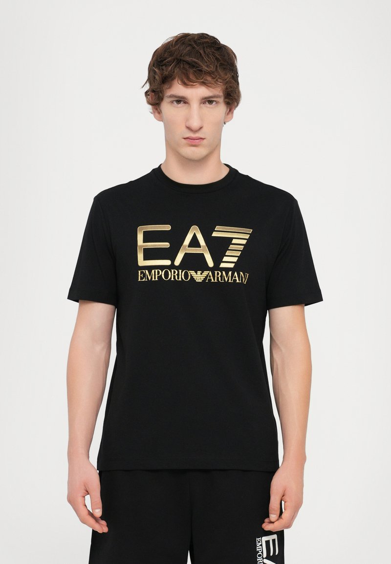 Black cotton t-shirt featuring a large gold printed logo "EA7 EMPORIO ARMANI" across the chest. Short sleeves, round neckline.