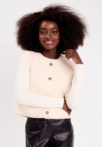 Femme souriante avec des cheveux bouclés volumineux portant un gilet crème à boutons sur un haut à manches longues et un pantalon en cuir noir, posant avec une main levée.